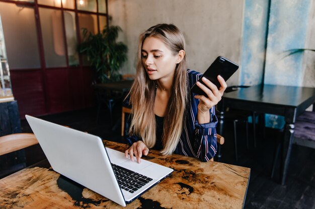 スマートフォンを手にノートパソコンで作業している素晴らしい白人の女の子。カフェに座っている魅力的な女性フリーランサー。