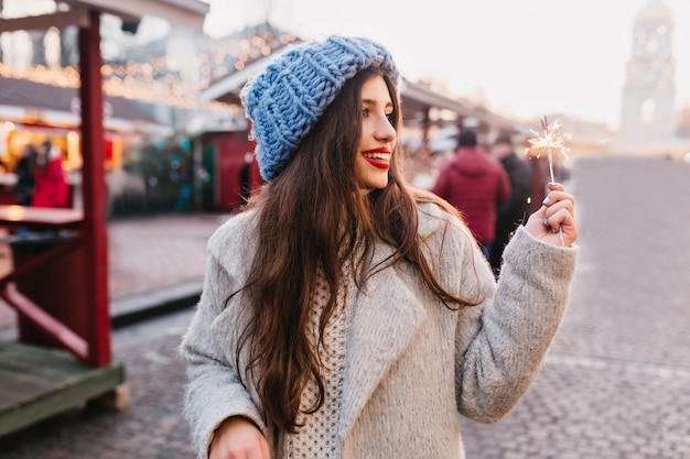 Amazing woman in gray coat and blue hat walking down the street with sparkler. Adorable female woman in winter outfit spending time outdoor and looking at Bengal light with smile.