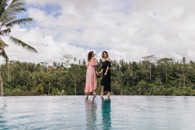 Amazing woman in long pink dress standing beside lake. Charming ladies holding hands near outdoor pool with forest