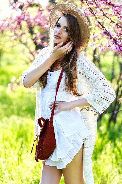 여름 시간에 햇볕이 잘 드는 정원에서 산책하는 모자에 긴 머리를 가진 흰색 빛 드레스에 놀라운 젊은 매력적인 여자. 피는 벚꽃, 밝은 색상, 카메라를 찾고, 세련된 민감한 모델, 휴식