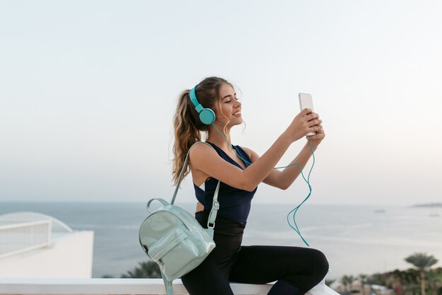 Amazing young attractive young woman in sportswear making selfie on phone in sunny morning on seafront. Resort, white colors, workout, cheerful mood, listening to music through headphones