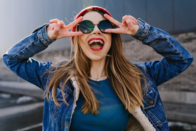 Amazing young lady in trendy attire laughing and making peace sign. Outdoor portrait of carefree positive girl in sunglasses having fun.