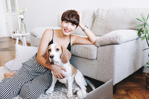 Free photo amazing young woman wears striped pants and wristwatch posing on the floor while playing with beagle dog