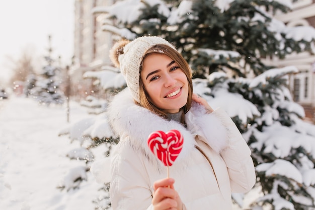 Удивительная молодая женщина в белой теплой одежде, вязаной шапке с розовым сердечком на палочке, веселится на улице. Привлекательная женщина, наслаждаясь зимним временем в городе.