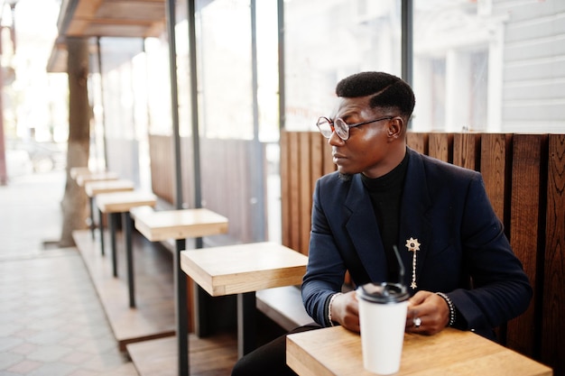 L'uomo afroamericano dall'aspetto sorprendente indossa un blazer blu con spilla a collo alto nero e occhiali in posa in strada ragazzo nero alla moda con una tazza di caffè che guarda il suo telefono