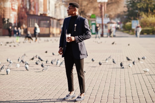 Amazingly looking african american man wear at blue blazer with brooch black turtleneck and glasses posed at street Fashionable black guy with cup of coffee