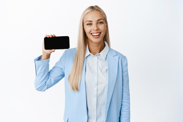 Амбициозная блондинка-корпоративная женщина в костюме, показывающая горизонтальный экран мобильного телефона, демонстрирующий белый фон веб-сайта компании-приложения