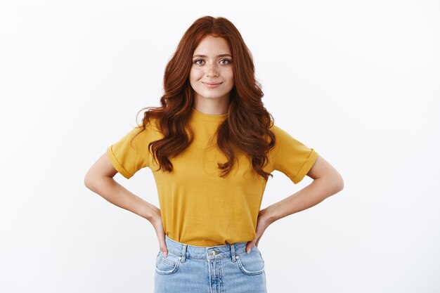 黄色のTシャツに赤い巻き毛の野心的で自信に満ちたかわいいヒップスターの女の子は、意欲的で自信に満ちた腰に手をつないで、明るくて断定的な笑顔、野心的なカメラ、白い壁を見つめています