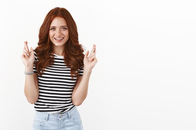 Ambitious confident ginger girl with freckles in striped tshirt cross fingers good luck smiling selfassured camera motivated win praying receive positive results making wish white background