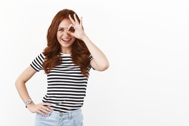 Ambitious goodlooking redhead flirty woman in striped tshirt long ginger curly hairstyle looking through fingers approvingly aim for success standing white background like offer