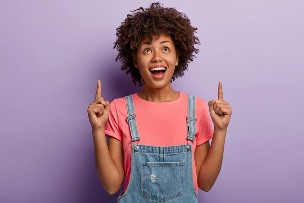 Ambitious pretty woman with curly hairstyle, points above, shows top copy space, smiles pleasantly, wears casual clothes, poses over purple wall