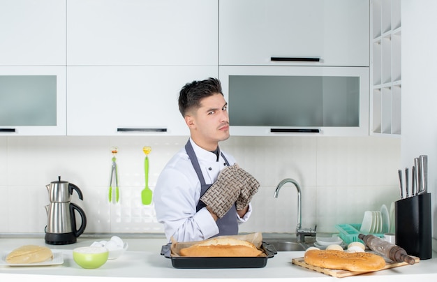 Free photo ambitious and proud commis chef in uniform wearing holder and freshly-baked bread in the white kitchen