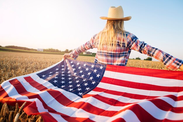 Американская женщина-фермер в повседневной одежде с распростертыми руками держит флаг США на пшеничном поле