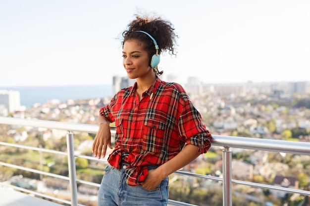 屋上にイヤホンでポーズをとって褐色肌のアメリカ人女性。都市景観の背景。