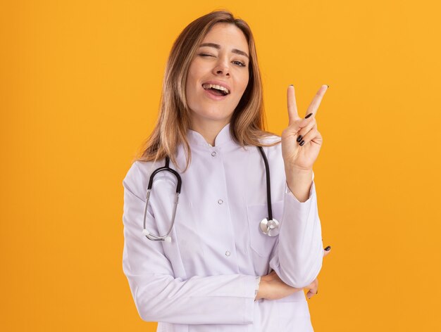 Amiling 노란색 벽에 고립 된 평화 제스처를 보여주는 청진 기 의료 가운을 입고 깜박이 젊은 여성 의사