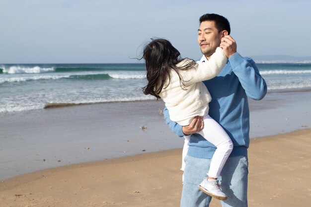 Amused Asian family walking together on beach. Father and daughter in casual clothes dancing near water and laughing. Togetherness, love, parenting concept