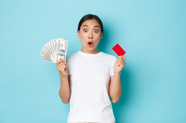 Amused asian girl in white casual t-shirt gasping, found out awesome prices, discount offers in shop, holding both credit card and cash, light-blue wall