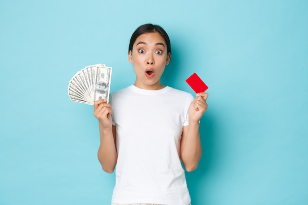 Забавная азиатская девушка в белой повседневной футболке, задыхаясь, узнала потрясающие цены, скидки в магазине, держит в руках кредитную карту и наличные, голубая стена