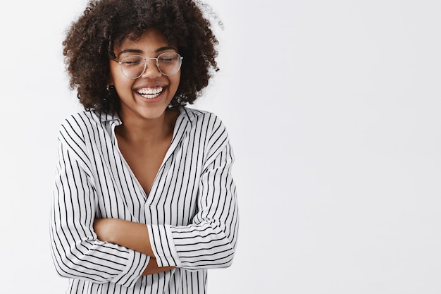 Amused and carefree attractive african american woman in striped blouse and glasses closing eyes laughing out loud and holding hands on chest closing eyes having fun