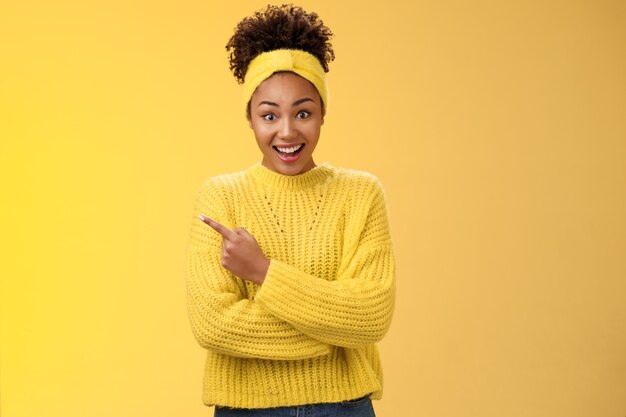 Amused charismatic smiling black cute girl in headband sweater widen eyes drop jaw astonished hear about awesome interesting new place talking friend standing yellow background pointing left amazed.