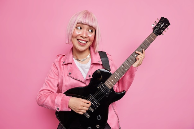 Amused cheerful pink haired rock star plays electric guitar being part of band dressed in jacket ready to jam on stage performs new song poses indoor has fun. Music entertainment hobby concept
