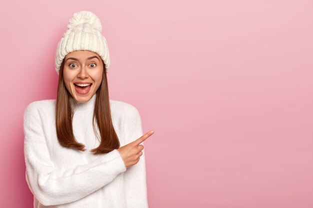 Amused cheerful woman points away with index finger, wears white hat and sweater, enjoys interesting scene, has long straight hair, grins and shows copy space