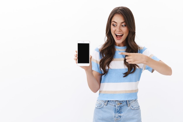Una joven alegre y divertida presenta una aplicación para teléfono inteligente, sostiene el teléfono móvil, señala y mira la pantalla, sonríe emocionada y recomienda una aplicación increíble, tienda en línea