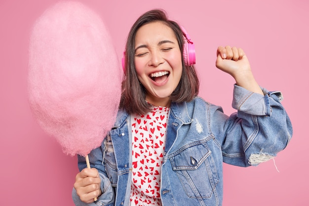 Amused dark haired teenage girl dances carefree moves with rhythm of music listens favorite song via headphones keeps arm raised holds cotton candy affords herself sugary dessert. Lifestyle.