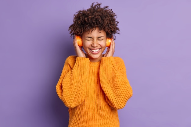 Amused dark skinned woman enjoys Xmas holiday playlist in headphones closes eyes and smiles toothily wears orange jumper poses over vivid purple background. Music lover indoor satisfied with sound