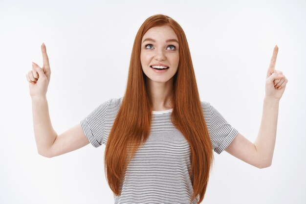 Amused dreamy thrilled joyful ginger girl blue eyes open mouth amazed look pointing index fingers up excited enjoy watching beautiful clouds fly sky standing daydreaming white wall