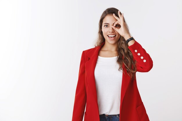 Amused enthusiastic dreamy gorgeous curly-haired caucasian girl wearing red jacket looking through okay gesture amazed excited open mouth astonished seeing opportunities, white wall