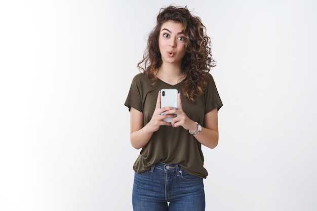 Amused good-looking young armenian curly-haired female impressed awesome cool smartphone camera taking photograph say wow fold lips surprised delighted capture good shots, standing white background