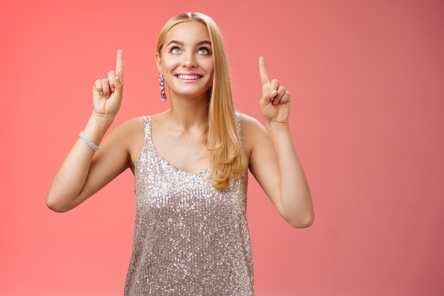 Amused gorgeous tender elegant european woman blond long hairstyle in silver glittering evening dress look pointing up curious gaze excitement see desired thing, standing red background joyful.