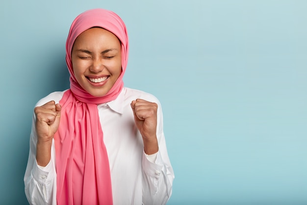 Free photo amused happy arabian woman raises clenched fists, gains goal successfully, keeps eyes shut