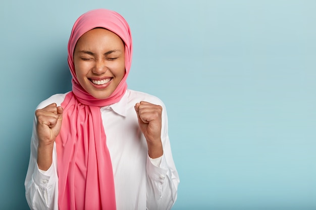 Foto gratuita la donna araba felice e divertita alza i pugni chiusi, guadagna l'obiettivo con successo, tiene gli occhi chiusi