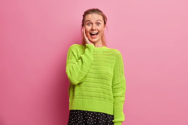 Amused happy European girl laughs out loudly, feels excited and very glad, keeps hand on cheek, stands upbeat, dressed in oversized knitted sweater