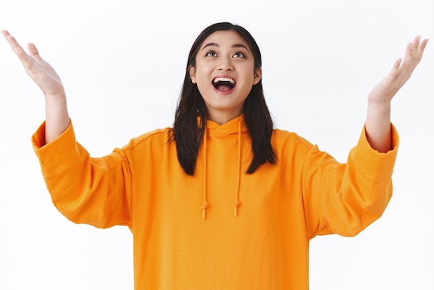 Free photo amused and happy triumphing young asian girl in orange hoodie raising hands up sky and smiling looking up thankful catching something or thanking heaven for dream come true