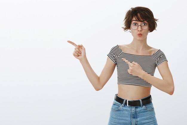 Amused hipster woman in glasses pointing fingers upper left corner at your logo or banner to find out details