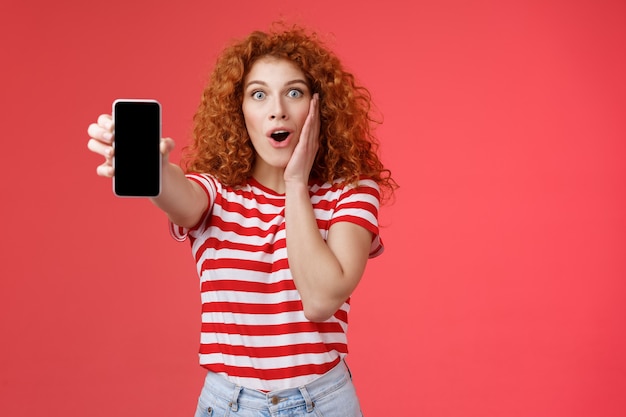 Amused impressed redhead good-looking curly-haired excited girl share awesome social media page show smartphone screen amazed touch cheek thrilled affected awesome app red background.