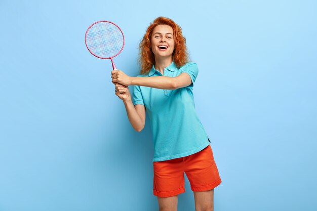 Amused playful red haired female likes playing tennis, holds racket