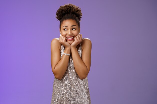 Amused silly cute black girl in silver dress anticipating exciting party dreamy gaze up touch cheeks smiling broadly optimistic waiting dream come true standing pleased touched blue background.