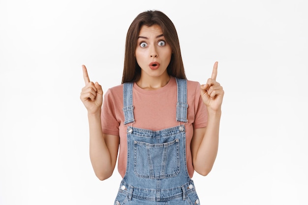 Amused surpirsed and impressed cute brunette female in overalls tshirt discuss amazing new product pointing fingers up folding lips and popping eyes astonished standing white background
