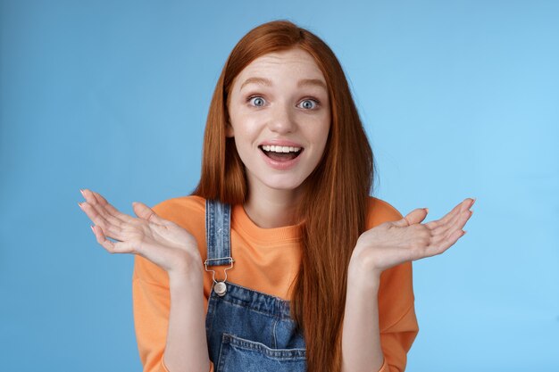 Amused surprised glad young redhead girl speechless happy see friend came back hometown wide eyes impressed grinning raise hands sideways full disbelief standing blue background joyful