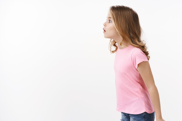 Amused wondered curious nosy little girl with blond hair stand in profile, open mouth fascinated attend amazing cool performance, look left copy space with interest and joy, pose white wall