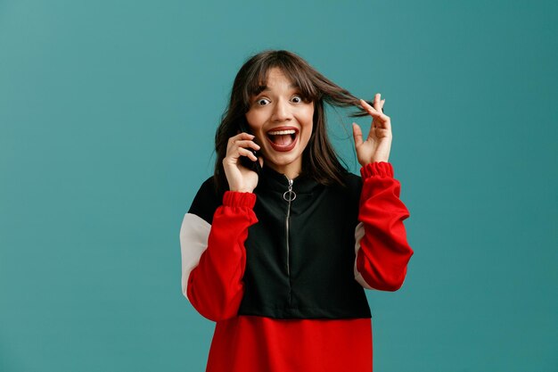 Amused young caucasian woman grabbing hair looking at camera while talking on phone isolated on blue background