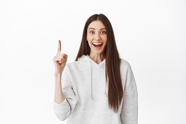 Amused young caucasian woman smiling in awe looking at camera impressed while pointing up at something cool showing awesome product best discounts standing over white background