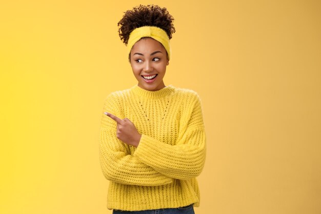 Веселая молодая милая африканская женщина-миллениал, выглядывающая глупо, указывая влево, широко улыбаясь, показывает любопытное интересное место, где наши друзья продвигают продукт с указанием рекламы, желтый фон.