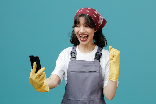 Удивленная молодая женщина-уборщица в униформе бандана и резиновых перчатках держит и смотрит на мобильный телефон, показывающий жест "да" на синем фоне