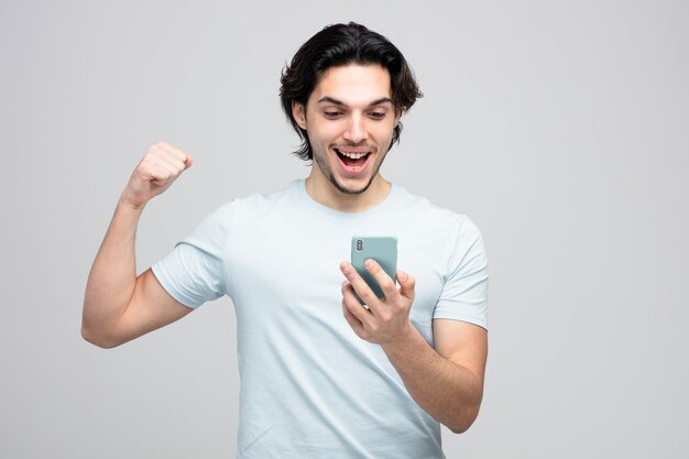удивленный молодой красивый мужчина держит и смотрит на мобильный телефон, показывая сильный жест на белом фоне