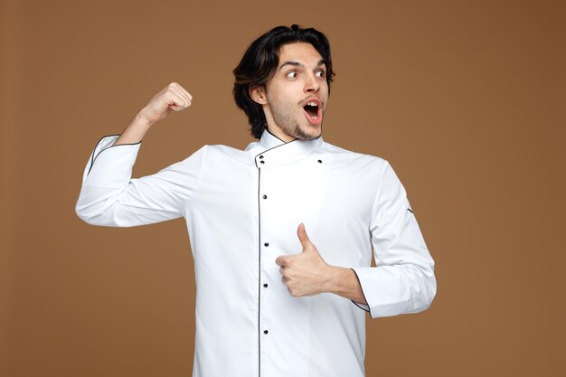 amused young male chef wearing uniform looking at side showing thumb up and strong gesture isolated on brown background